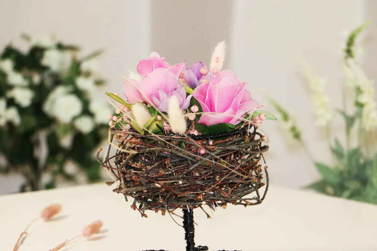 gavebokse Et delikat fuglerede laget av tynne kvister står på sort stativ; i redet ligger rosa silkeblomster og hvite harehalestrå mot uskarp, lys bakgrunn.